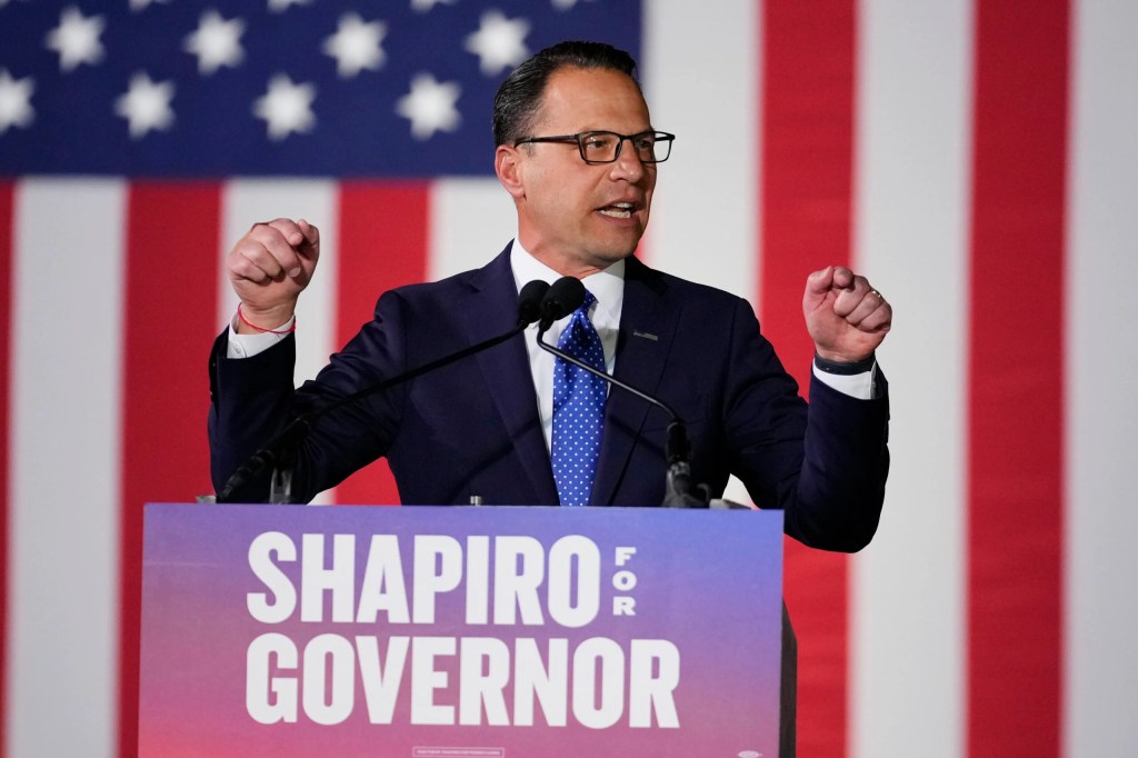 Josh Shapiro at a podium speaking into a microphone, an American flag in the background.