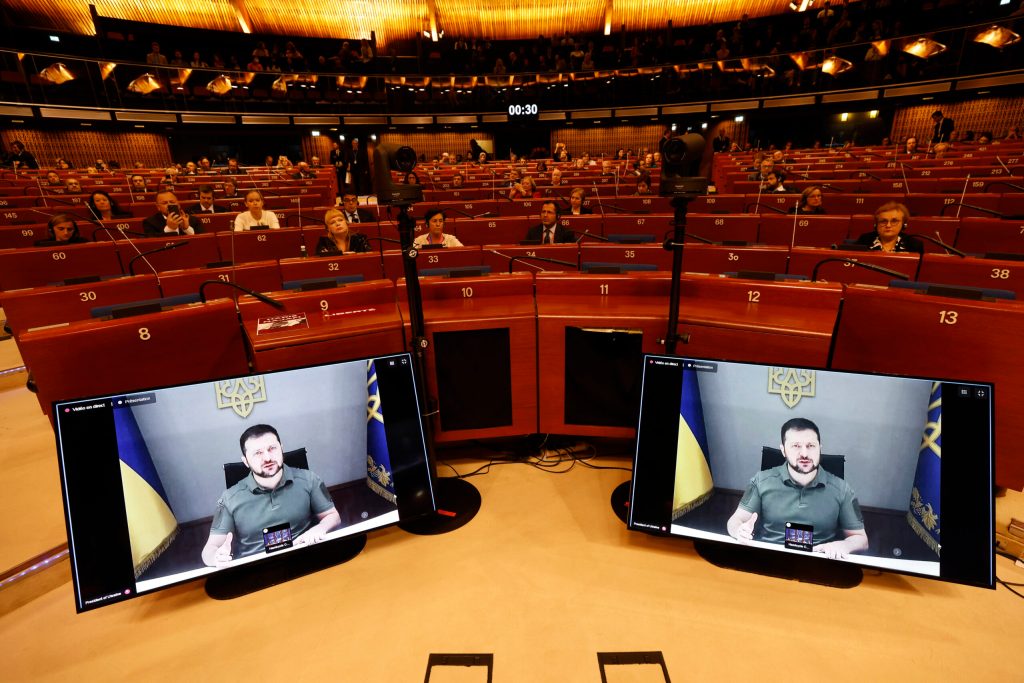 Ukrainian President Volodymyr Zelenskyy shown on video conference on two computer monitors facing the camera. The background shows members of the European council attending the meeting.