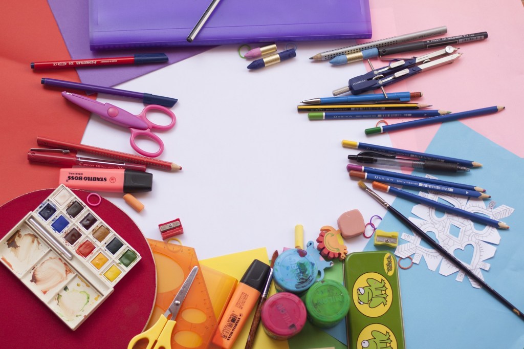School supplies, including pens, pencils, paintbrushes, watercolor paint, highlighters, and construction paper. All are arranged in a circle according to color, creating a rainbow.