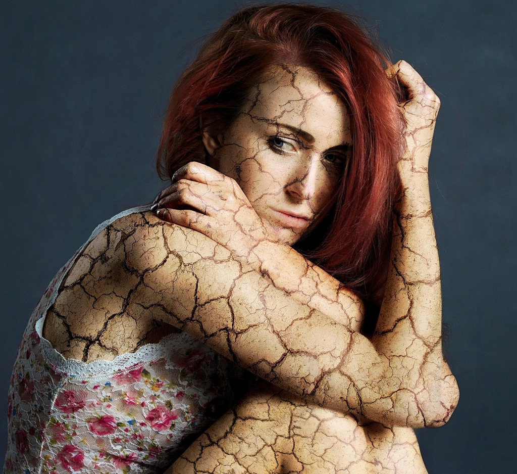 A woman with red hair sitting with her knees drawn up, elbows resting on them. Cracks have been Photoshopped onto her skin.
