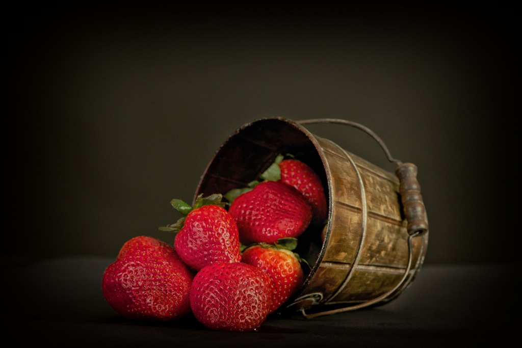 A small bucket turned on its side with strawberries spilling out on a black background.