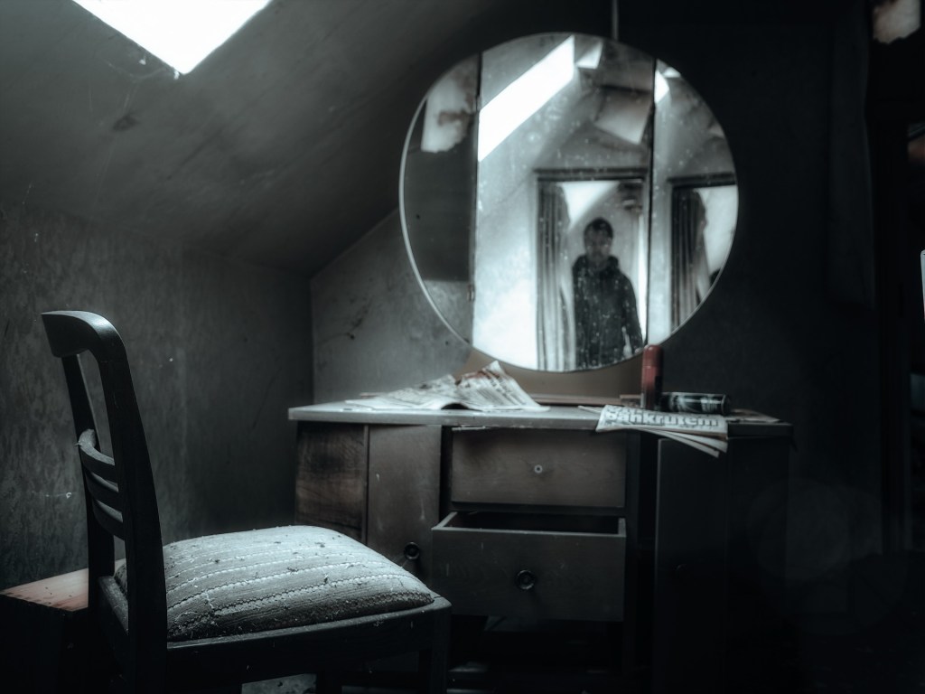 A chair in front of a vanity mirror, both spotlit in a dark room. A figure can be seen in the mirror in the doorway of the room.