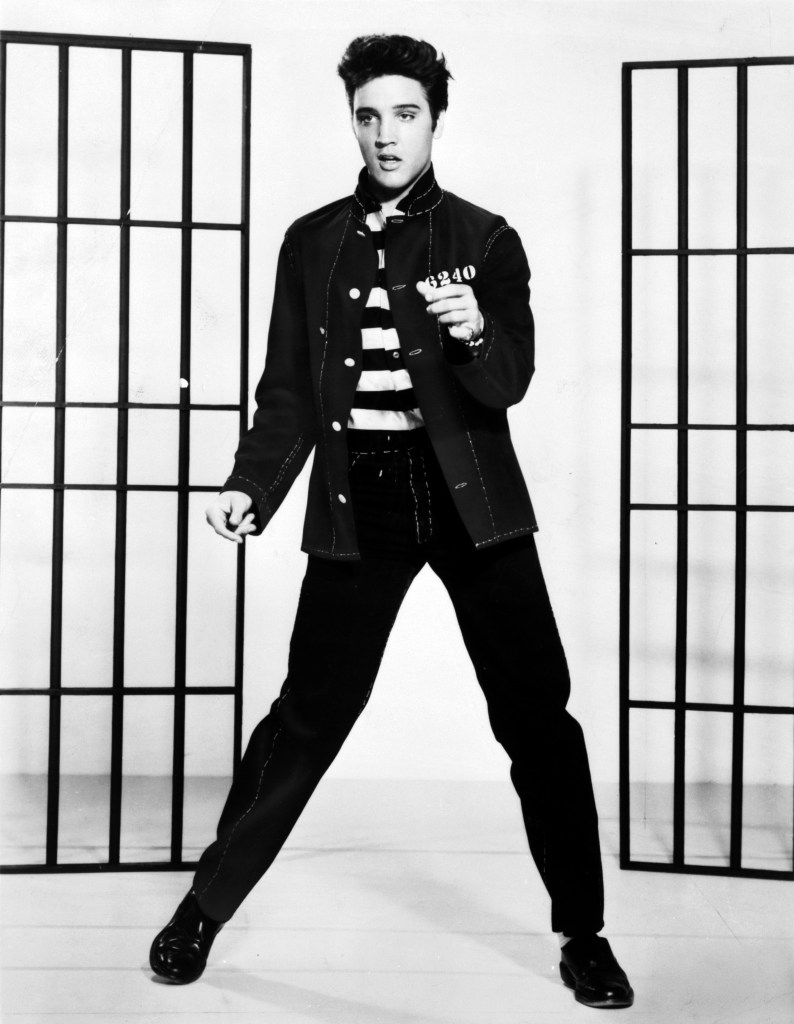 Elvis Presley posing in a white and black striped shirt, black jacket, and pants in a promo shot for "Jailhouse Rock."