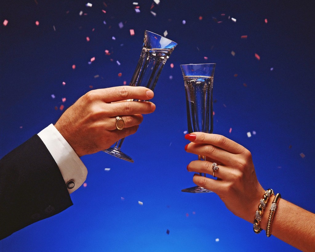 Two hands toasting with flutes of champagne on a dark blue background, red confetti falling down.