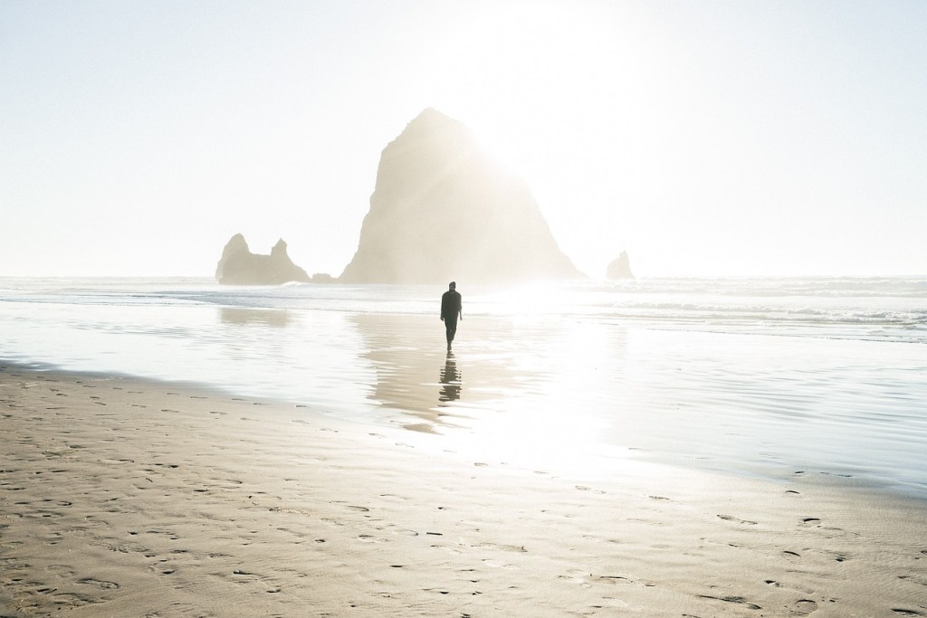 A lone figure walking on a tranquil beach, the sun overexposing the seascape around them.