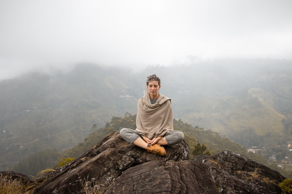 A woman sitting in the lotus position on top of a rock with her eyes closed. Fog obscures the background behind her.