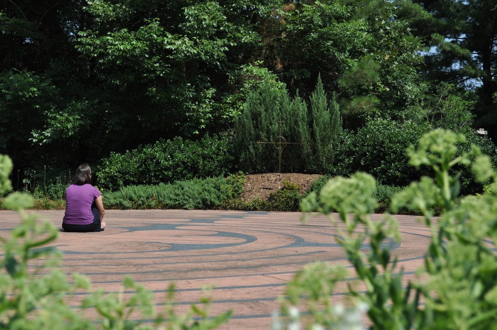 A woman sitting in the center of a stone labyrinth laid into the ground. She is contemplating a cross while surrounded by greenery.